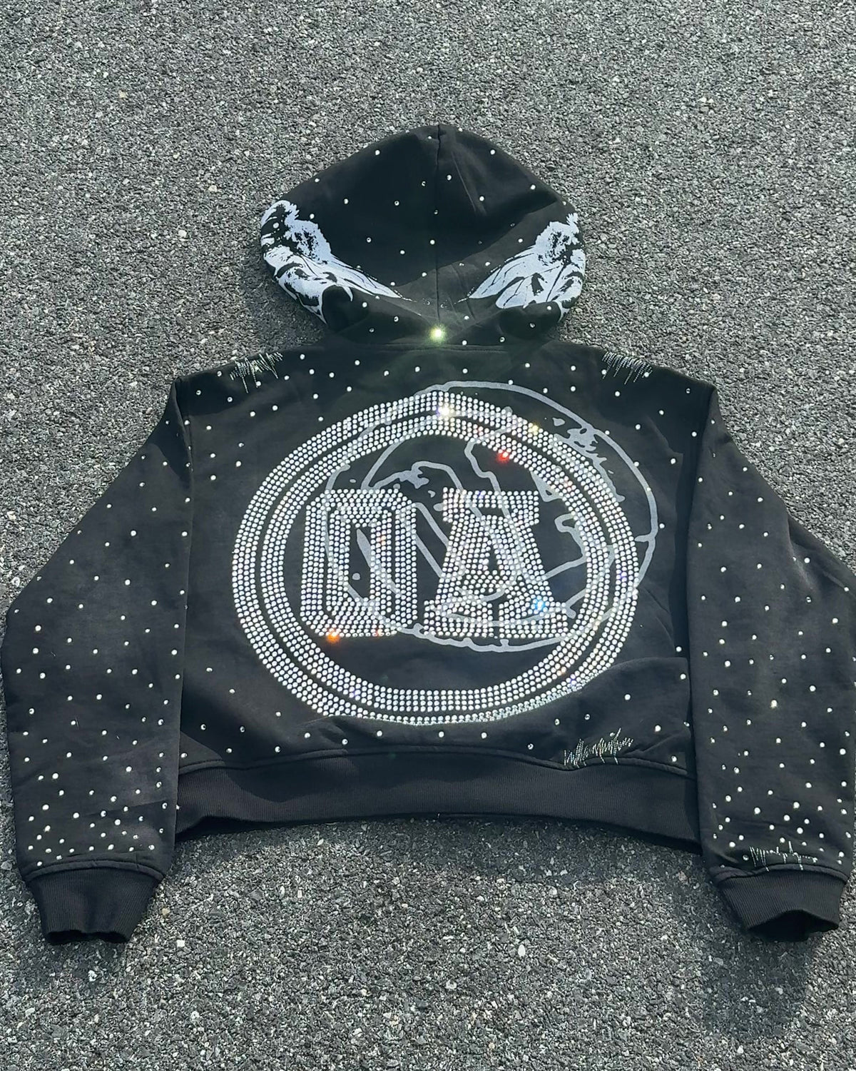 OUT THE MUD zip-up hoodie in black laid flat on the street, back with the rhinestone OA crest