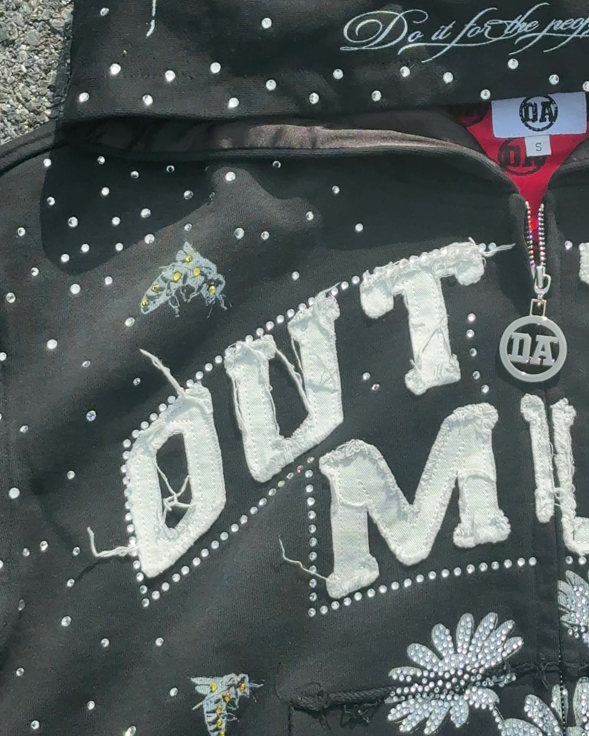 Close-up of the OUT THE MUD lettering, rhinestone daisies, OA zipper pull and the red lining at the neck